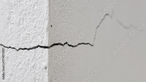 Close-up of a distinct diagonal crack in the corner of a rough white painted concrete or stucco wal, showing damage and decay.