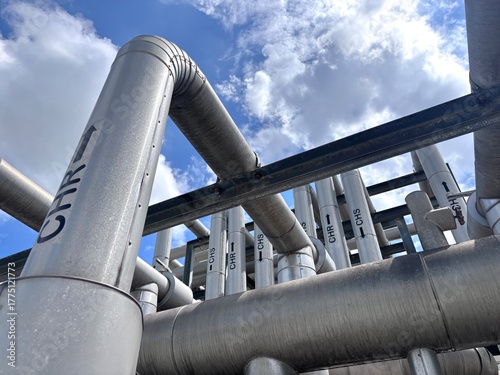 Low-angle abstract view of an industrial pipe network, featuring insulated metal pipes with identification labels and flow direction arrows against a bright blue sky and white clouds.