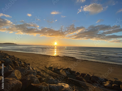 Golden Sunset Over Ocean Horizon with Sandy Beach, Foreground Boulders, and Coastal Waves
