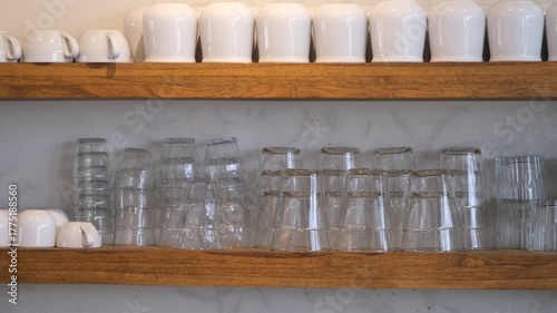 Smooth panning video showing neatly arranged white ceramic mugs and transparent glass cups placed upside down on wooden shelves against a marble wall.
