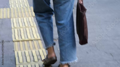 Handheld, slightly shaky close-up video of a person walking on a sidewalk with yellow tactile paving, wearing jeans and sandals while holding a brown tote bag.  