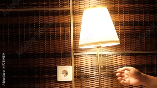 a hand reaching out to turn off a glowing table lamp against a woven rattan wall with a nearby power socket.  
