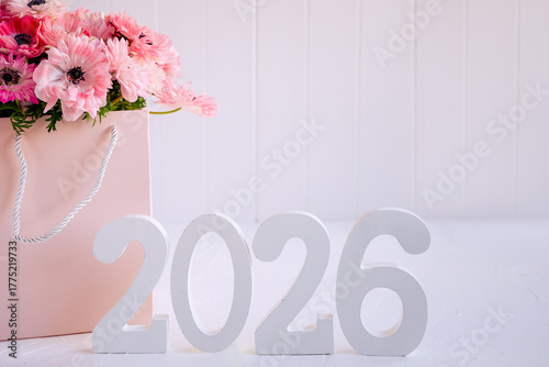 White wooden numbers forming 2026 standing on a light surface with a soft pink gift bag holding blooming flowers, symbolizing new beginnings and celebration