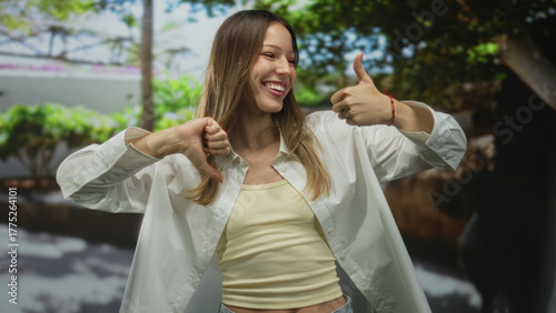Woman in white shirt and yellow tank top gives thumbs up and thumbs down on street while smiling and frowning, showing mixed gestures; ambivalence indecision choice.