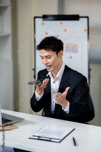 A businessman is talking to an AI via smartphone on his desk in an office.
