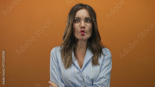Young hispanic woman with glasses puckers lips and crosses arms in a brightly lit orange studio; playful humor.