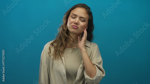 Woman feeling discomfort touching her cheek isolated against a solid blue background wearing casual attire with a neutral expression conveying distress and discomfort.