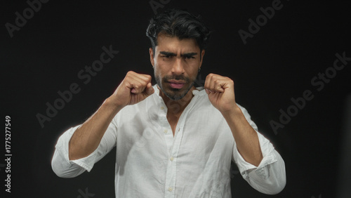 Young hispanic man rubbing his eyes in a black studio setting, wearing a white shirt and a tired expression; fatigue.