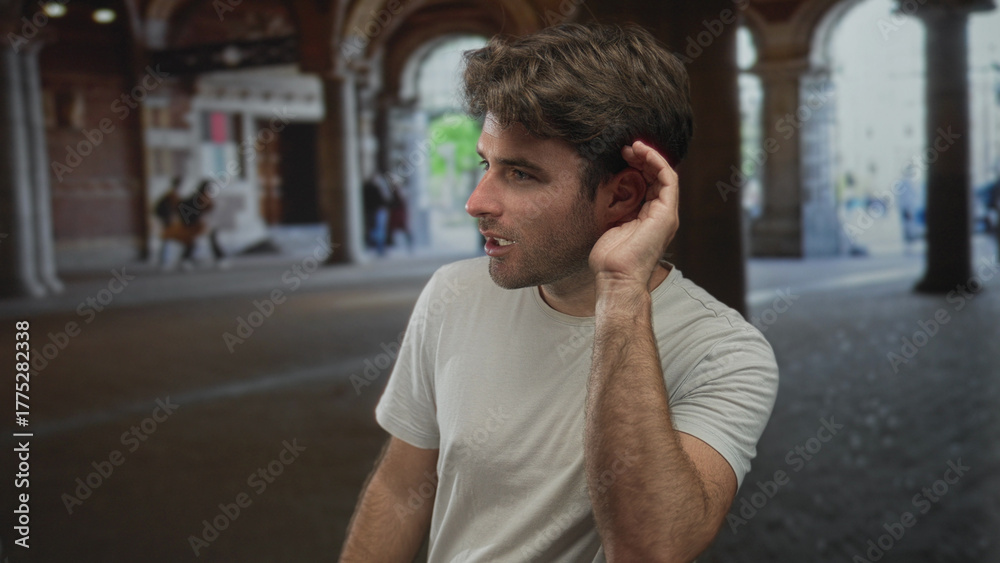 Fototapeta premium Man cups ear with hand to listen under arched city street arcade, smiling and leaning forward; curiosity attentiveness.