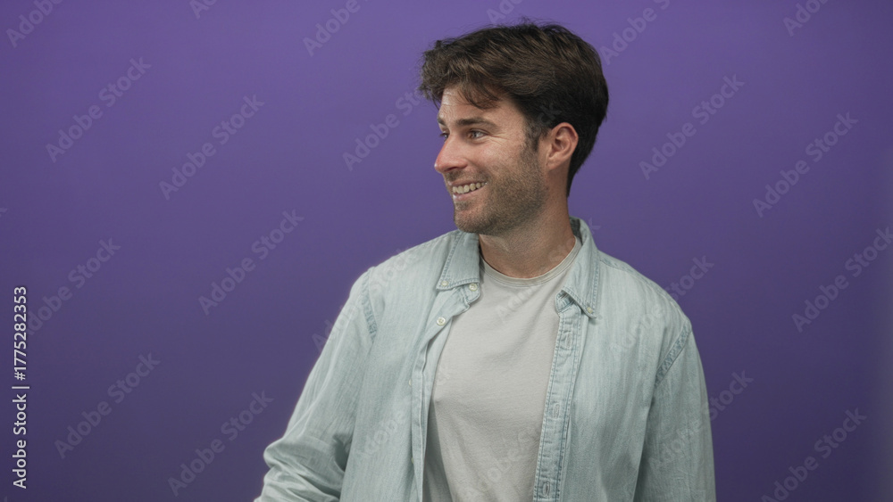 Obraz premium Man points thumb to side in a purple studio wearing light denim shirt and white tee, smiling while looking off camera; friendly casual.