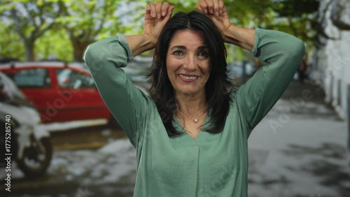 Slika na platnu Hispanic middle aged woman smiles and lifts hands behind head in bunny ears gesture on urban street; playful joy fun whimsy