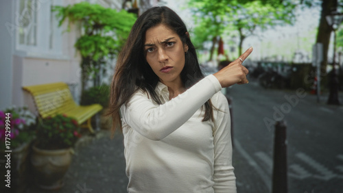 Young hispanic woman finger pointing to object on urban street with stern frown and forward posture; anger accusation.