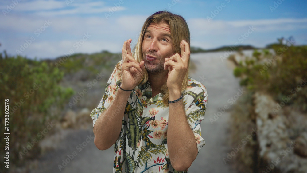 Obraz premium Blond man with long hair holds crossed fingers with both hands on a gravel path in a green park under blue sky; hope luck.