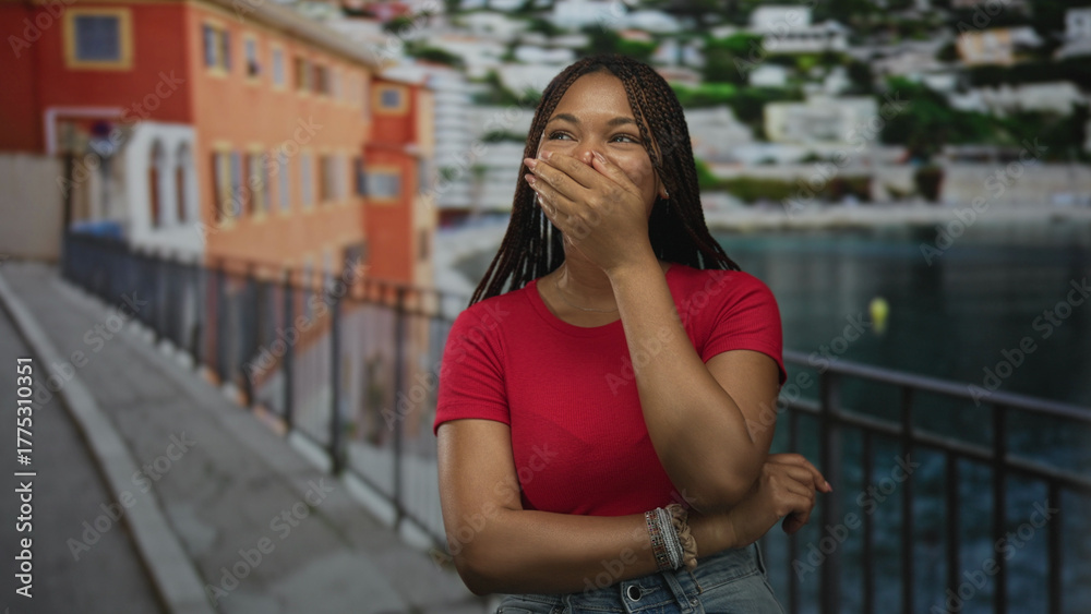 Fototapeta premium Woman with hand covering mouth on a seaside street promenade near railing and water, braided hair and red tshirt; surprise wonder.
