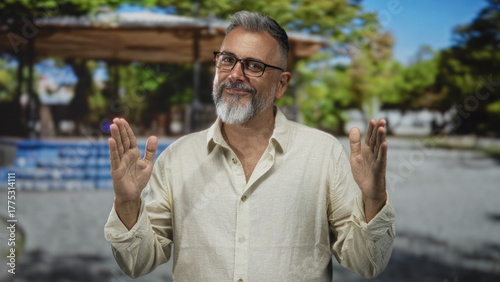 Man with eyeglasses and gray beard holds hands apart with palms visible on street near a pavilion and trees, wearing linen shirt and open collar; confidence.