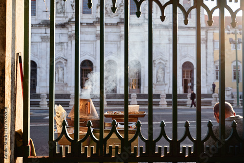 A street vendor selling food behind a decorative green gate, with smoke rising from the cooking area. In the background, a historic building is visible, and a person is seated nearby.