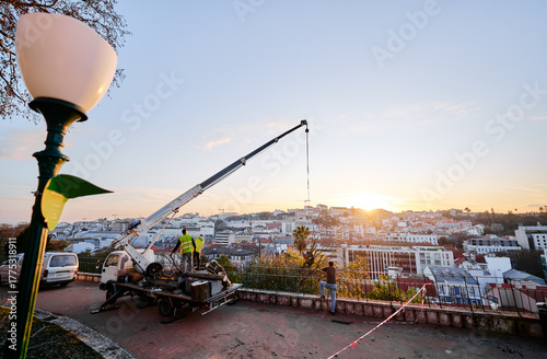 Workers operating a crane on a hilltop overlooking a cityscape at sunset.
