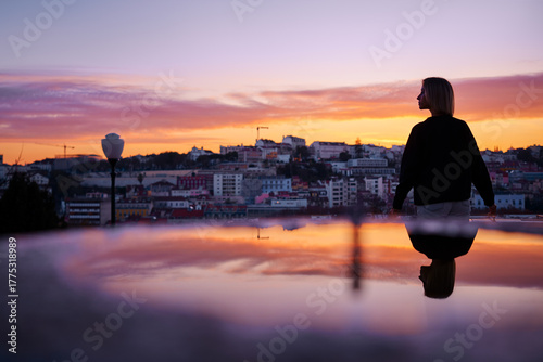 Silhouette of a person standing by a reflective surface at sunset, overlooking a cityscape with colorful buildings and a dramatic sky.