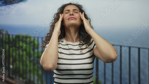 Schilderij op canvas Woman presses temples with hands on street railing beside the sea under bright daylight; stress and fatigue