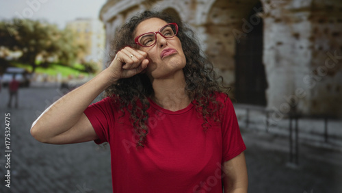 Young hispanic woman outdoors rubs eyes for crying at ancient roman coliseum building; sadness longing.
