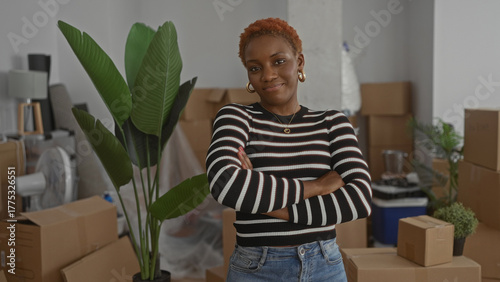 Obraz na plátně Young african american woman with crossed arms stands amid cardboard moving boxes and a tall green houseplant in a bright building interior; confidence renewal