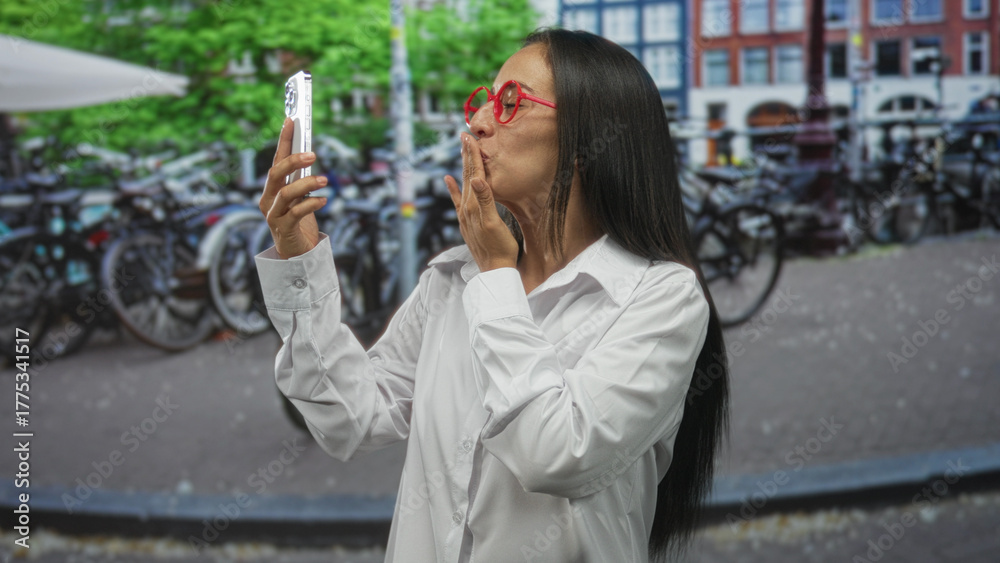 Fototapeta premium Woman holding smartphone and blowing a kiss on an amsterdam street while wearing red glasses and white shirt; affection connection.