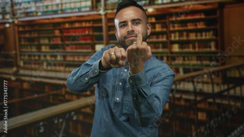 Tableau sur toile Young man in denim shirt shows middle finger gesture toward camera in a library building filled with books; defiance rebellion