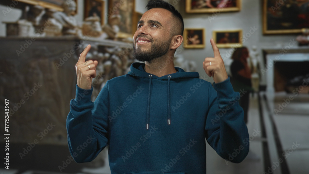 Obraz premium Man pointing both index fingers upward inside museum building with framed paintings and sculptures; playful curiosity.