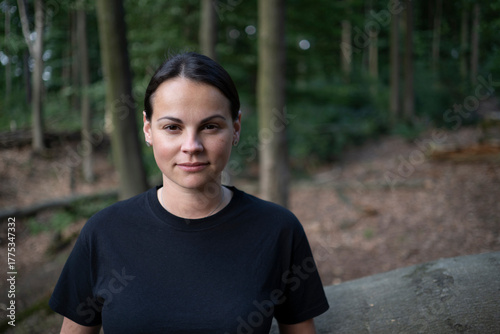 A young woman in a black T-shirt standing in front of a fallen tree trunk in the forest, waist-up, with a slight smile. Soft natural light, blurred trees in the background