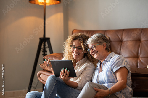 Fotografie Portrait of happy mature blonde curly woman sitting on the floor close to her elderly mother sharing together social news on digital tablet