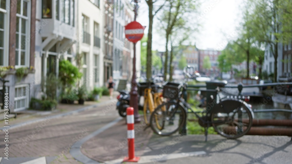 Obraz premium Defocused view of bicycles and red sign in a sunny amsterdam street with trees and canal, capturing urban tranquility and charm of a typical netherlands city scene.