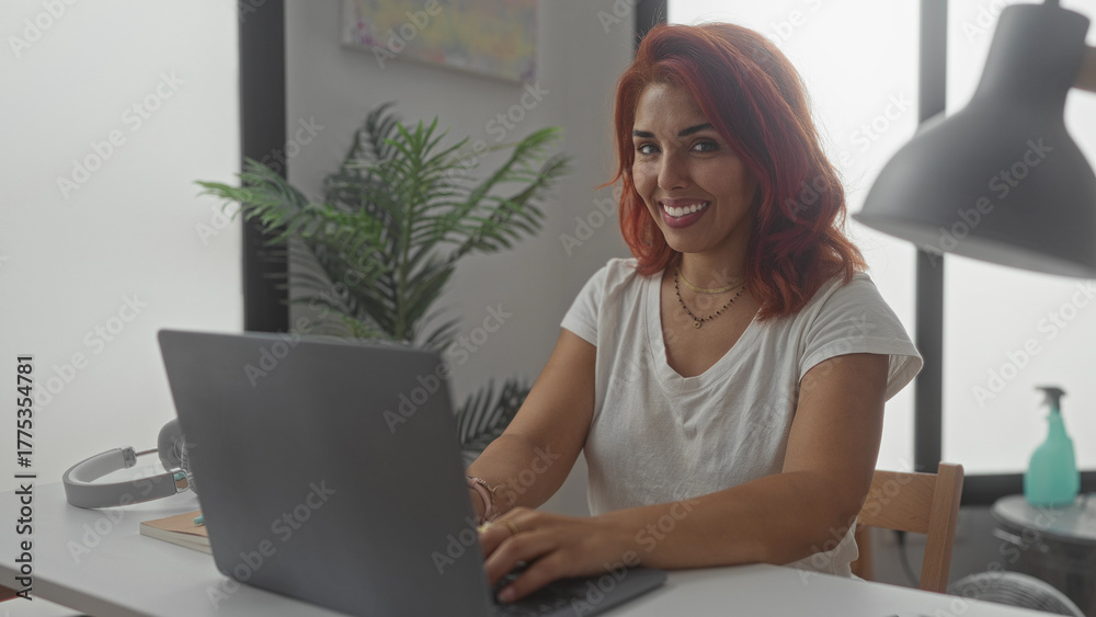 Fototapeta premium Woman typing on laptop in building home office at desk with headphones and plant visible; remote work focus.