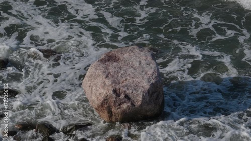 Sea waves breaking on coastal rock with dramatic white water spray and foam texture. Scenic overhead view of natural ocean landscape with a stone boulder in turbulent, flowing surf water.