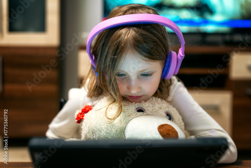 Little girl wearing headphones watching tablet screen and hugging teddy bear, concept of child technology use and digital dependence