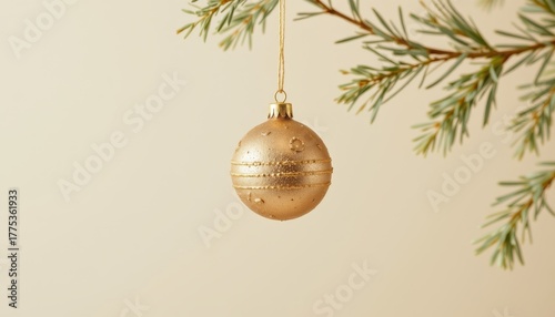 Single textured gold Christmas ornament hangs from a blurred green pine branch against a beige background