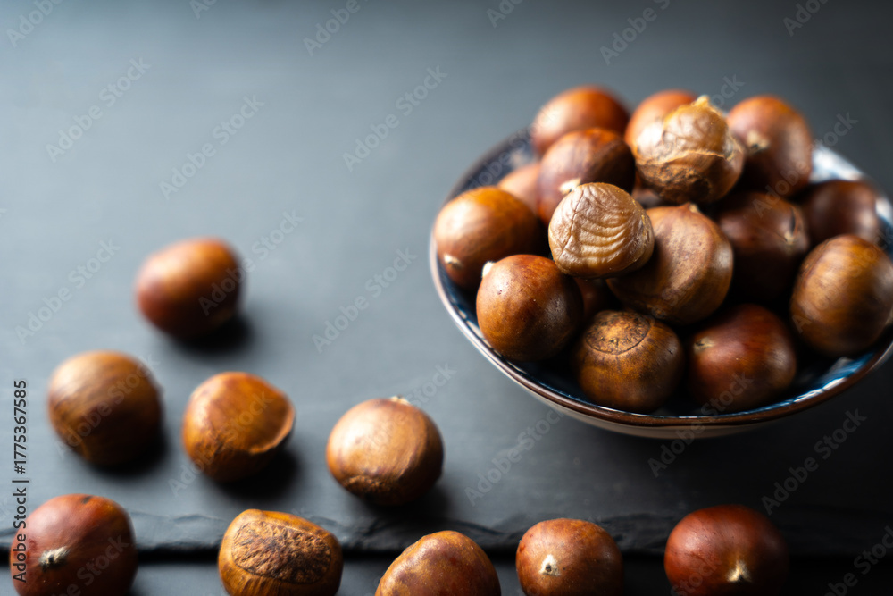 Fototapeta premium Fresh raw chestnuts in a bowl on black background, healthy organic autumn food, natural seasonal nut rich in flavor and nutrition, perfect ingredient for cooking and roasting.