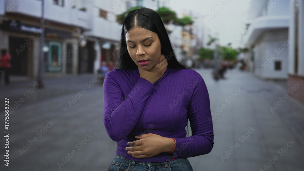 Fototapeta premium Young latin woman with long black hair clutching neck on city street wearing purple top and blue jeans; discomfort.
