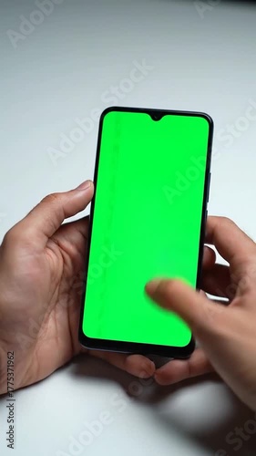 Human hands holding a smartphone with a vibrant green screen, centered on a light surface