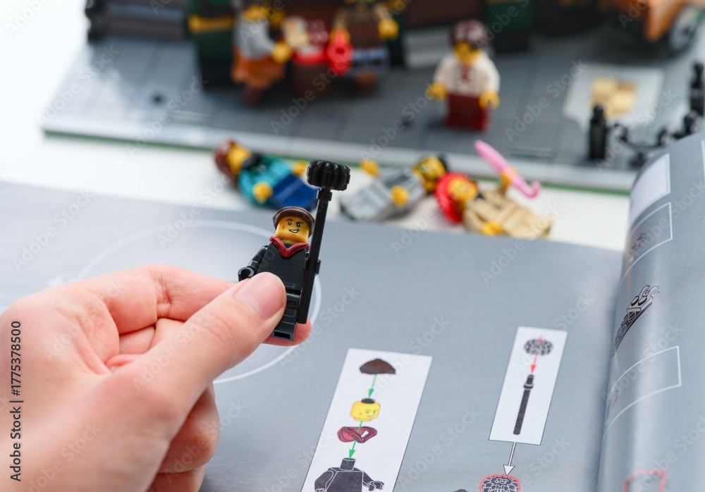 Fototapeta premium Tambov, Russian Federation - June 28, 2025 Person hand holding a built Lego chimney sweep minifigure on an instruction booklet. Some minifigures and Lego set in the background
