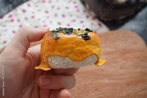 a hand holding 1 on 1 replica of japanese onigiri topped with mentai sauce and seaweed on wooden background