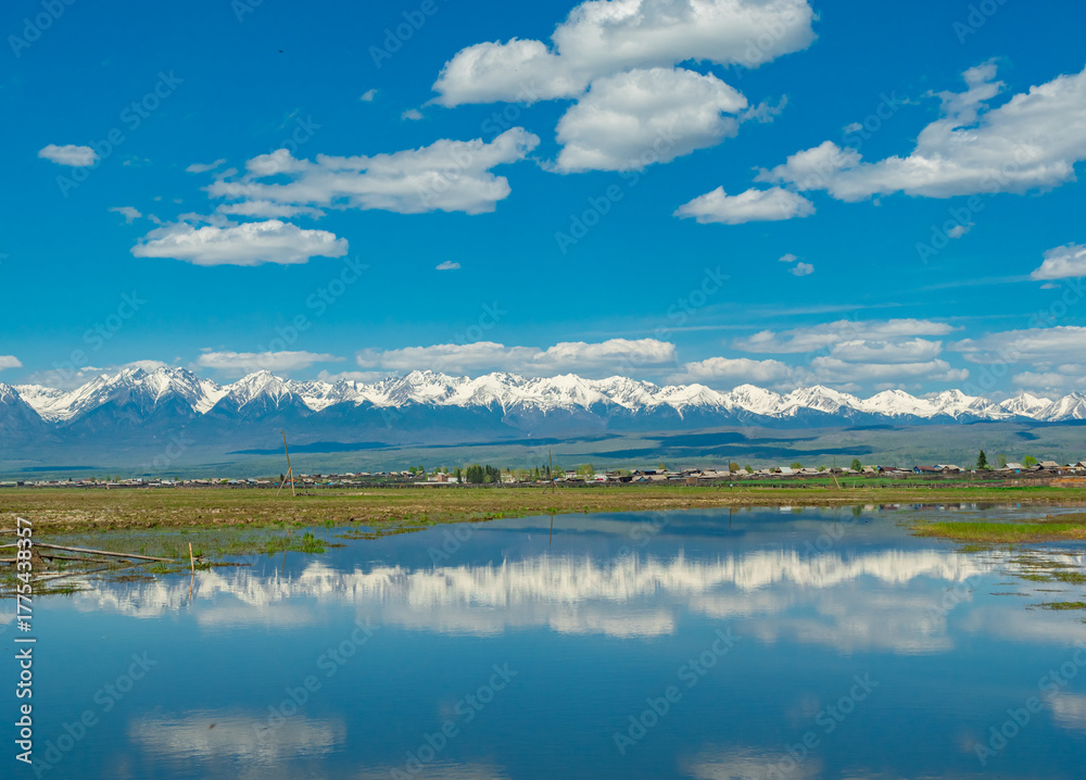 Obraz premium Majestic snow-capped mountains reflect in serene lake under bright blue sky with fluffy clouds in summer