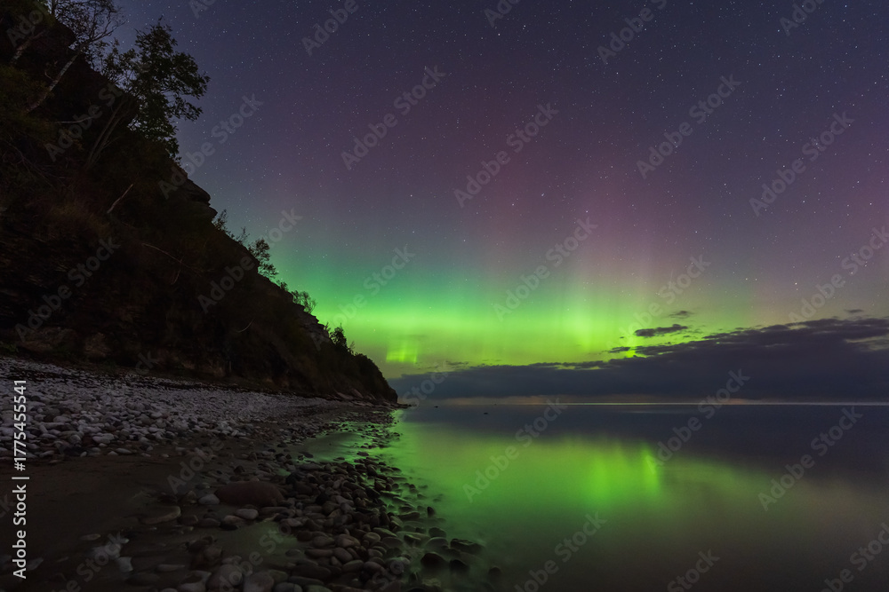 Naklejka premium Stunning green and purple Aurora Borealis (Northern Lights) reflecting in calm water on the rocky coast of Estonia.