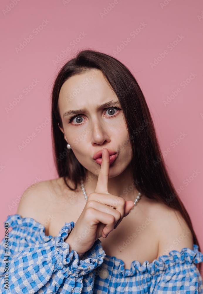 Fototapeta premium Woman in blue checkered top making a shushing gesture against a pink background.