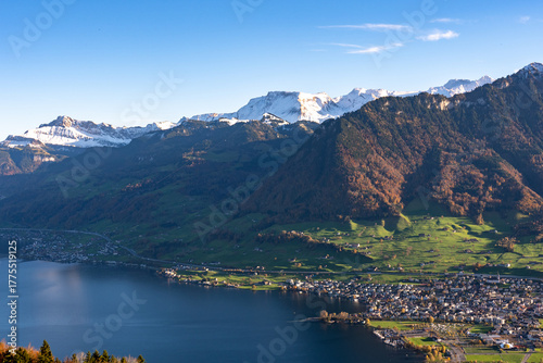 A high-angle view captures a densely settled lakeside town nestled between dark blue water and steep, sun-drenched alpine slopes with snow-capped peaks against a clear sky.