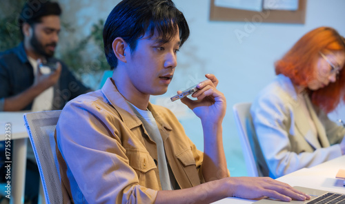 Asian man using AI voice assistant on smartphone in office, interacting with virtual assistant app for productivity, smart technology, and team collaboration.