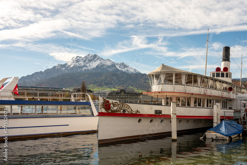 Historic paddle steamer and modern tourist boat docked, with Mount Pilatus' snowy peaks dominating the urban backdrop under a blue sky.