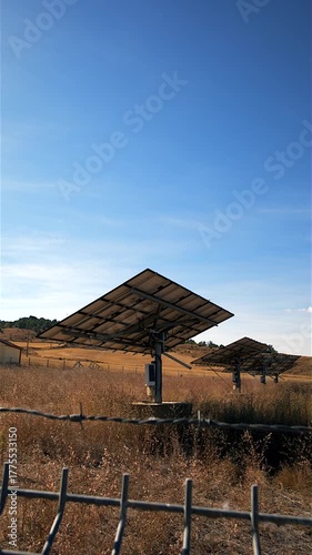 Solar panels are set up in a parched field, enclosed by a wire mesh fence, producing eco friendly energy from sunlight on a bright and sunny day in a rural setting under a clear blue sky