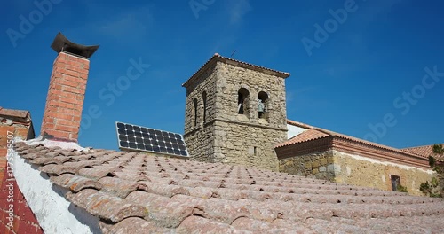 Contemporary photovoltaic solar panels mounted on aged tiled roof of historic stone church, blending traditional architecture and renewable energy under clear blue sky, europe