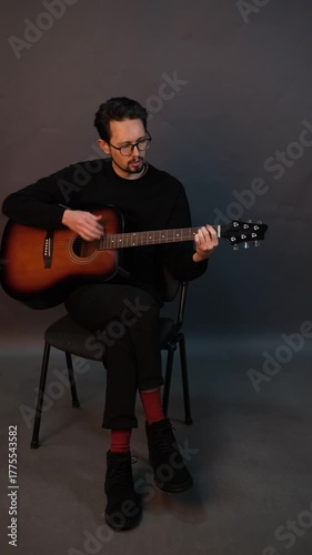 A handsome young brunet man playing guitar while sitting on a chair in a dark studio

