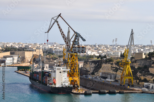 Valletta, Malta - September 24, 2025: Cargo cranes and large ship docked near sandstone fortifications and boats in the harbor.